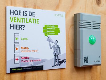 iotta-klimaatsensor met groene stoplichtindicatie naast een instructiebord over ventileren, in een leslokaal