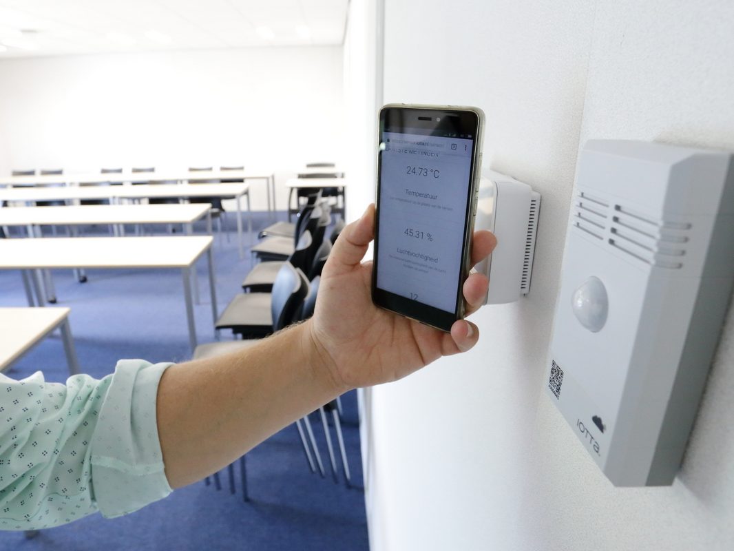 Hand die met een smartphone de QR-code van een wandgemonteerde iotta-sensor scant en de actuele meetwaarden op het scherm ziet